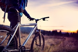 A low wide angle view of an unrecognizable man looking toward the setting sun in the horizon, holding his bicycle as he prepares for the final run of the day.