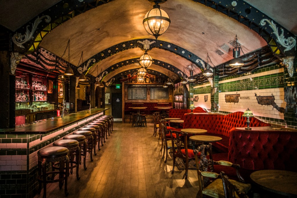 This is a photo of the Whitechapel Bar. It features wooden floors, a bar on the left with tiling and red sofas on the right. The ceiling is dome-shaped.