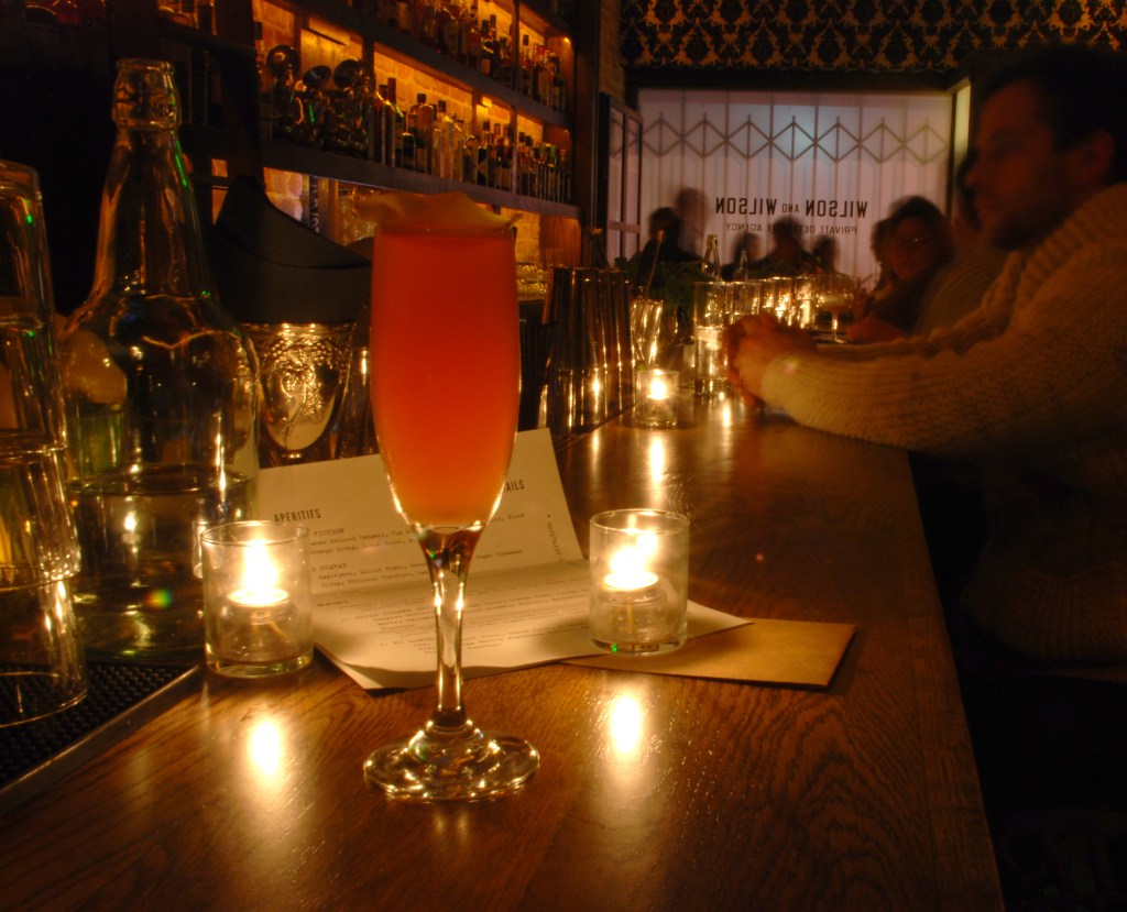 This is a photo of a pink drink sitting on top of a bar. The bar is dark with candle lighting.