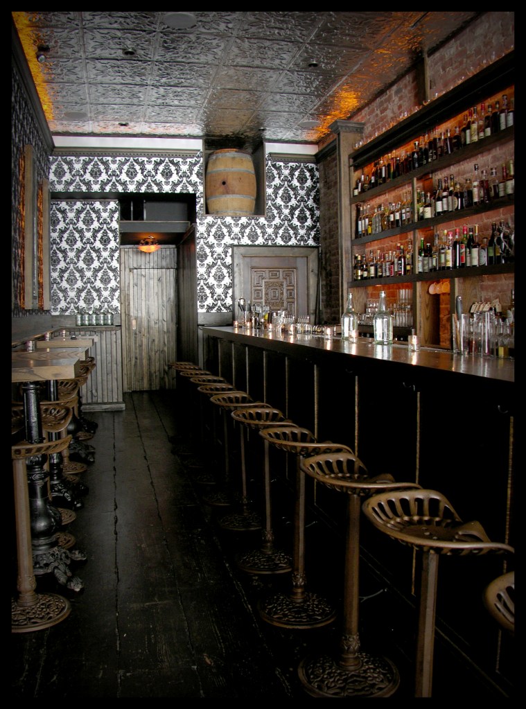 This is a photo of the Wilson & Wilson bar. It's long and narrow with wooden stools on the right and gray wall paper on the back wall.